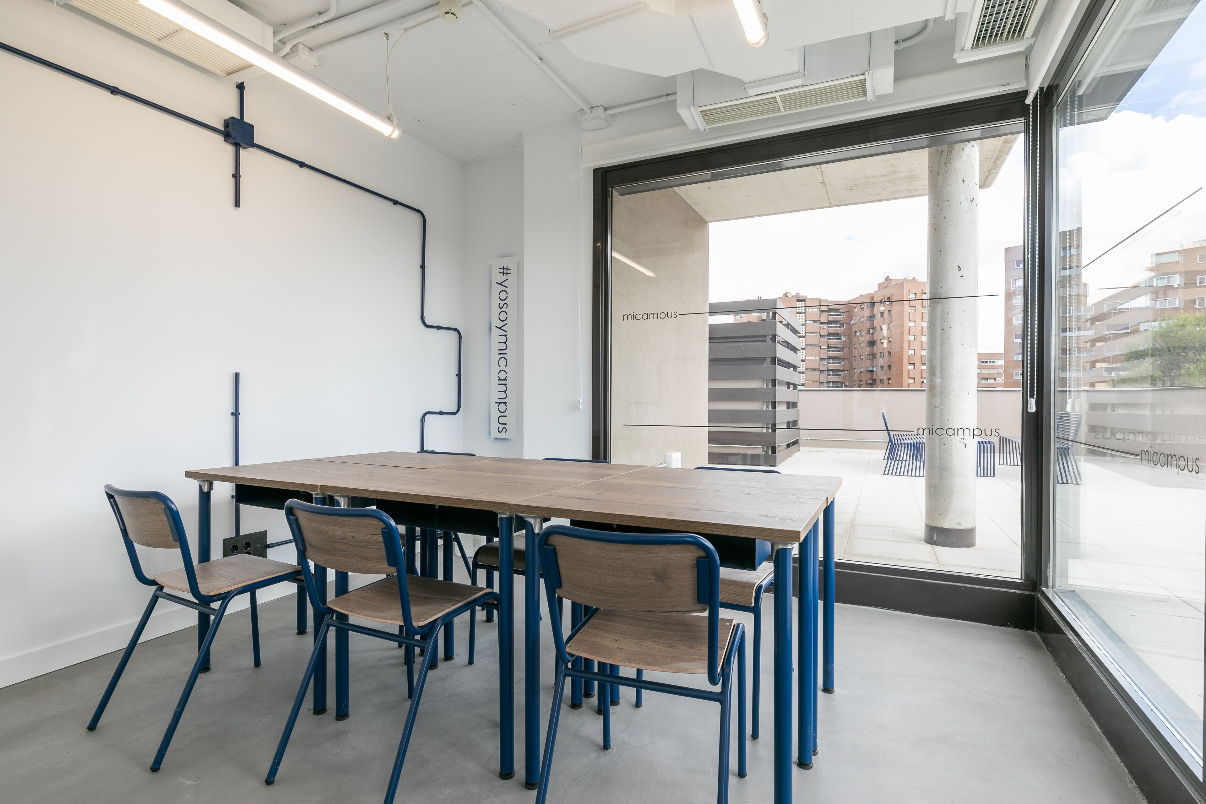 A conference room with a long table and six chairs, large windows with a view of buildings, and a white wall with a black pipe.