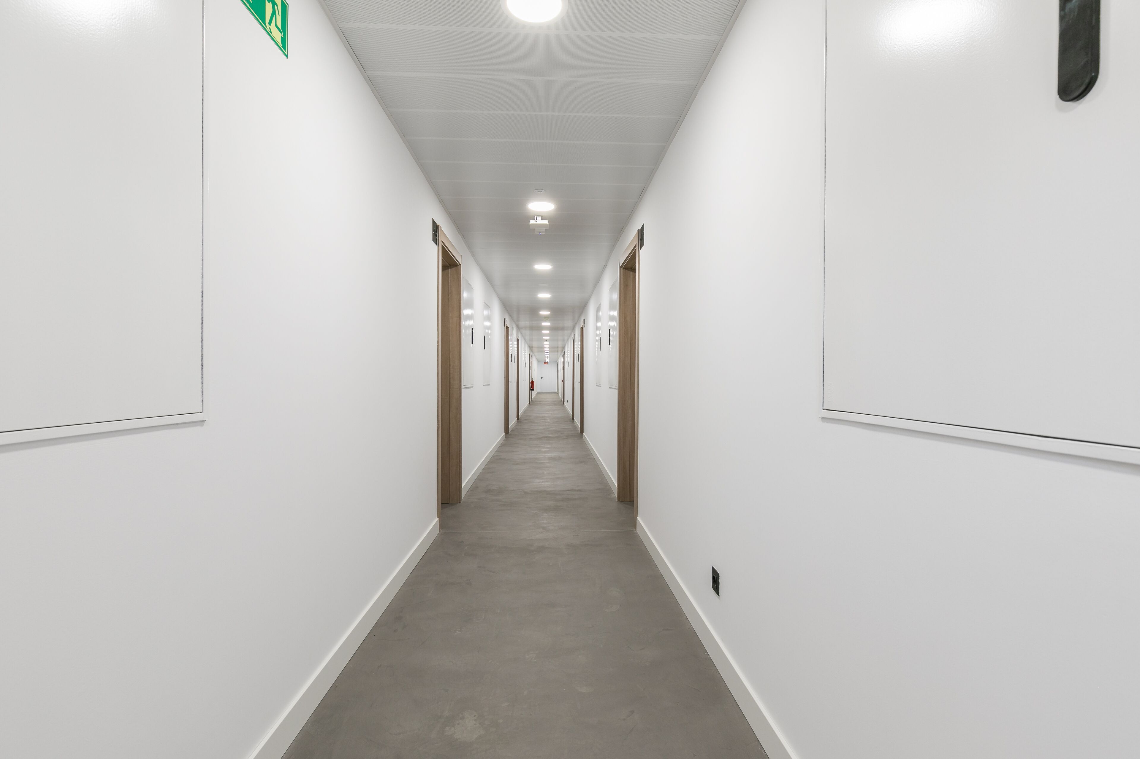 Un largo pasillo estrecho con paredes blancas y suelo gris, que conduce a varias puertas a ambos lados.