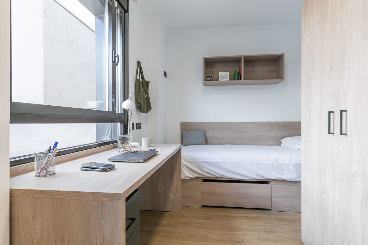 A compact bedroom with a wooden desk, a bed with a headboard, a shelf with books, and a window with a view.