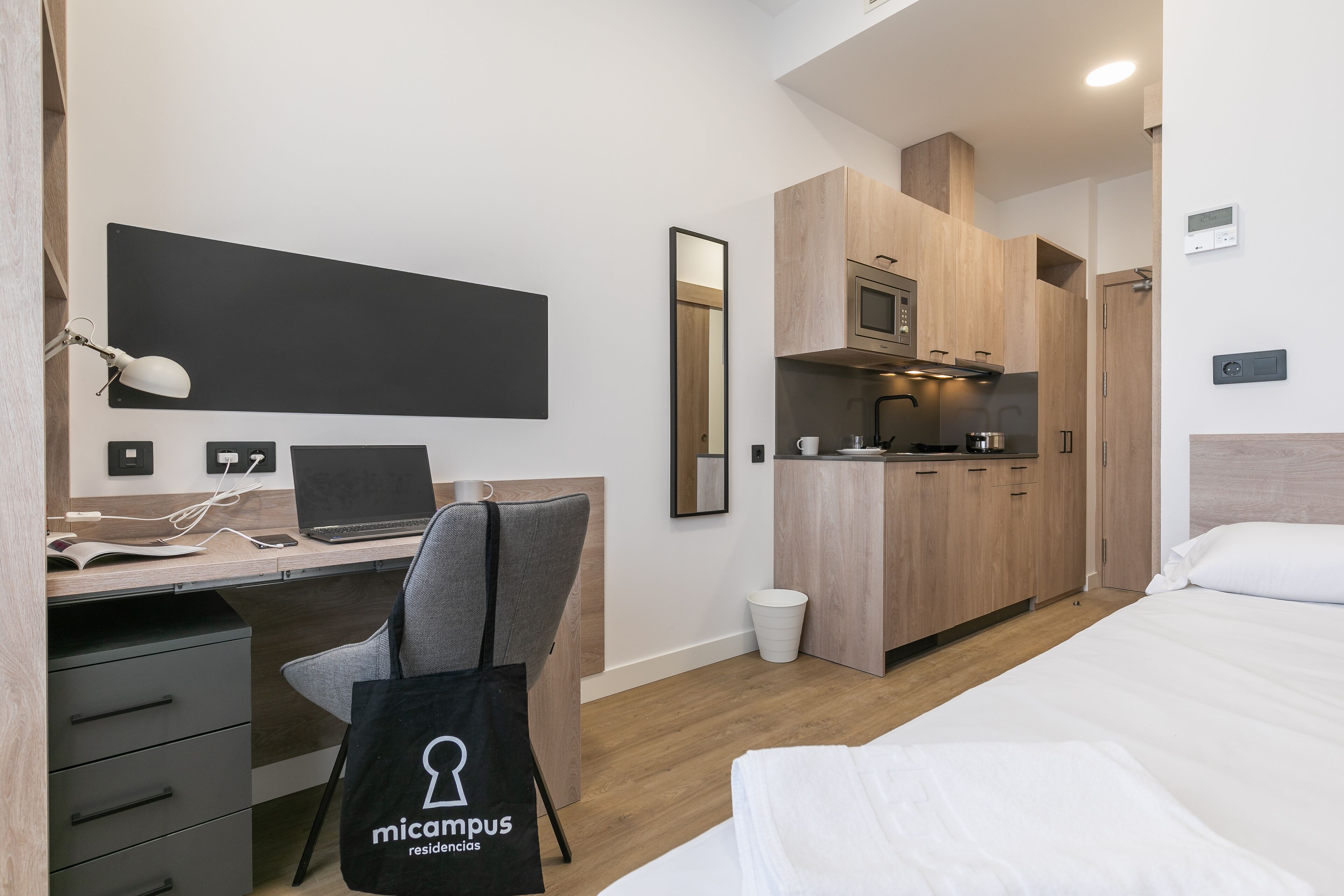 A modern hotel room with a bed, desk, and kitchenette.