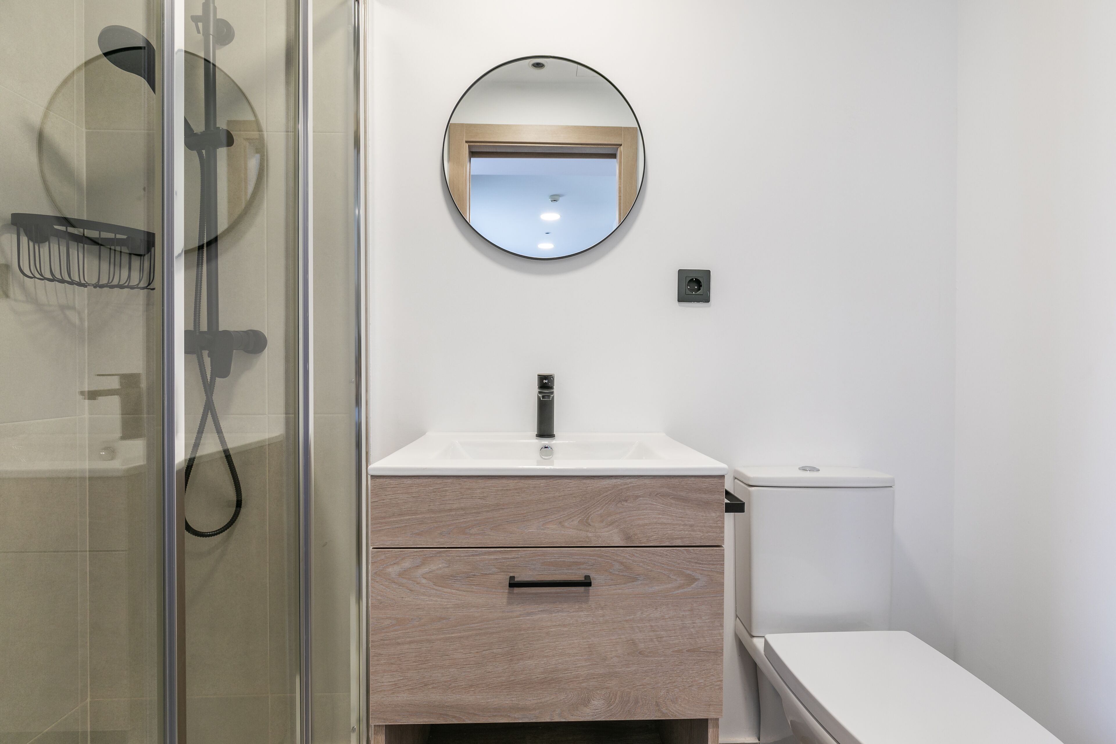 A modern bathroom with a glass shower enclosure, a white sink with a wooden vanity, and a round mirror above the sink.