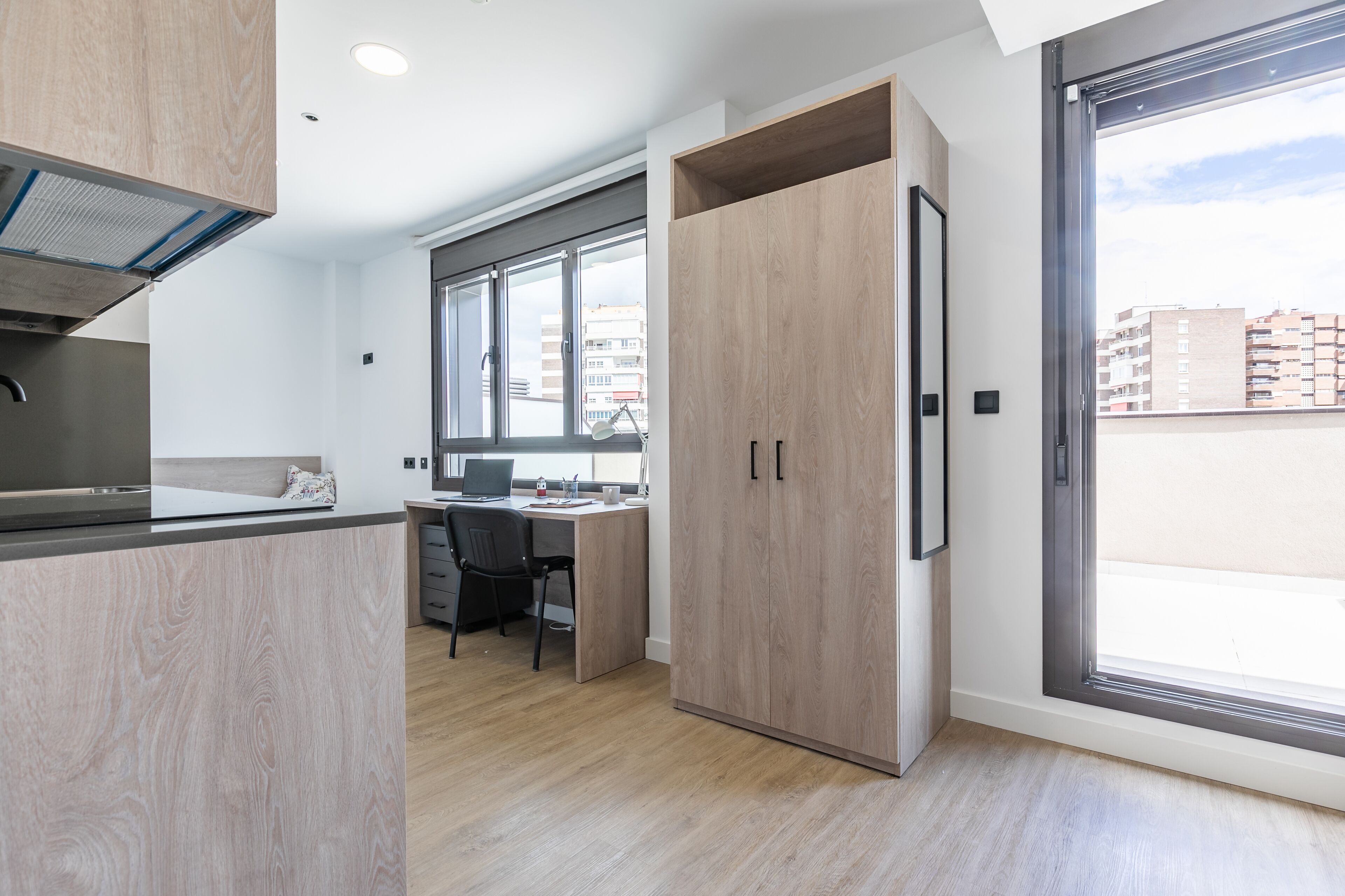 A modern kitchen with a built-in stove, a dining area with a table and chairs, and a large sliding door leading to a balcony with a city view.