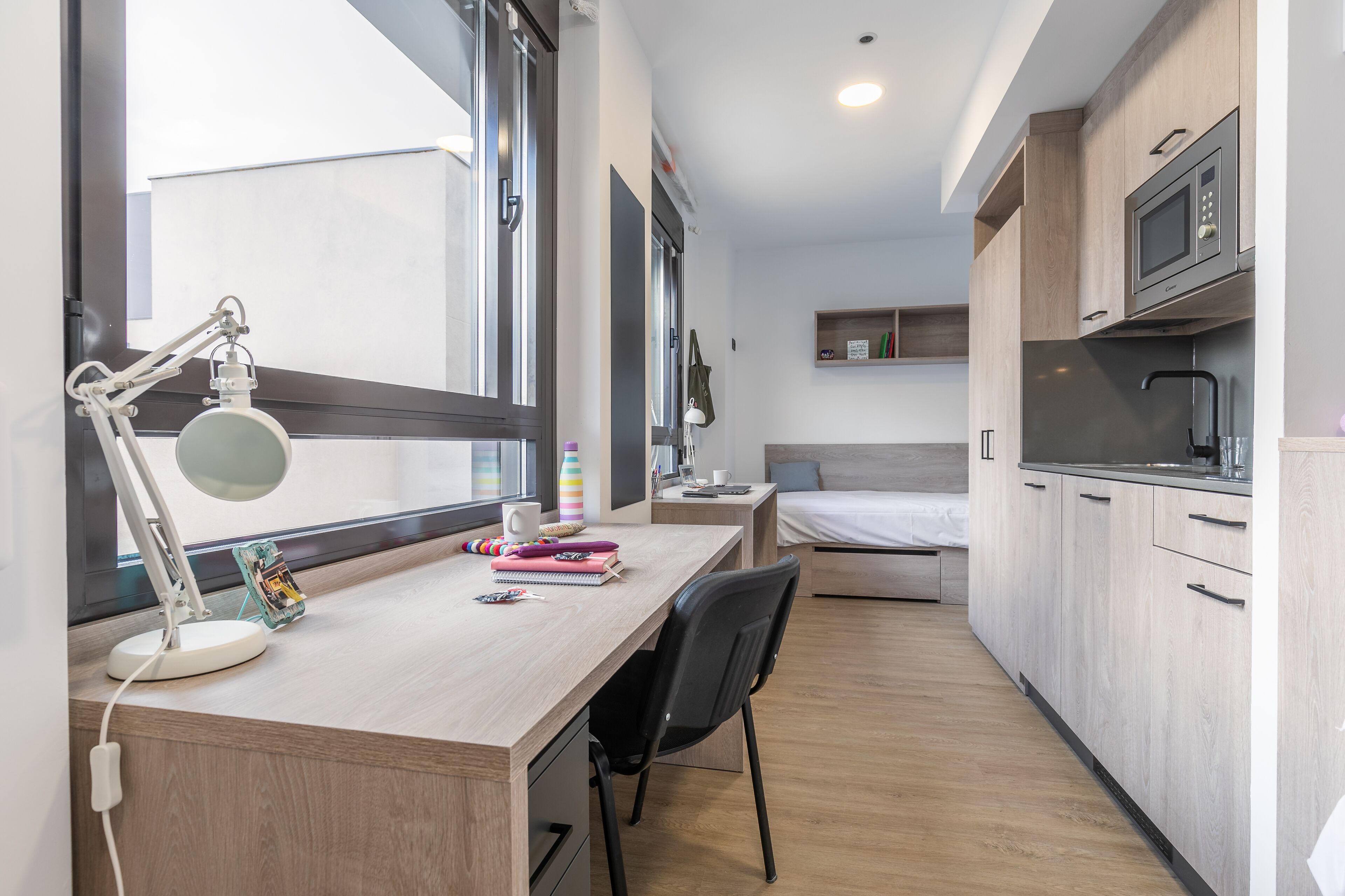 Una habitación compacta y moderna con escritorio de madera, una silla, una cama y una kitchenette.