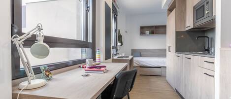 Una habitación compacta y moderna con escritorio de madera, una silla, una cama y una kitchenette.