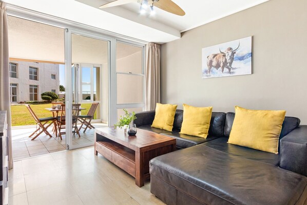 A modern living room with a leather sofa, wooden coffee table, and yellow cushions. There is a sliding glass door leading to an outdoor area with a patio table and chairs.