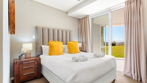 A bedroom with a large bed, a wooden nightstand, a lamp, and a view of the outdoors.