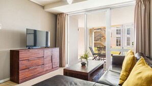 A modern living room with a flat-screen TV on a wooden cabinet, a sofa with yellow cushions, and a sliding glass door leading to a balcony with outdoor furniture.
