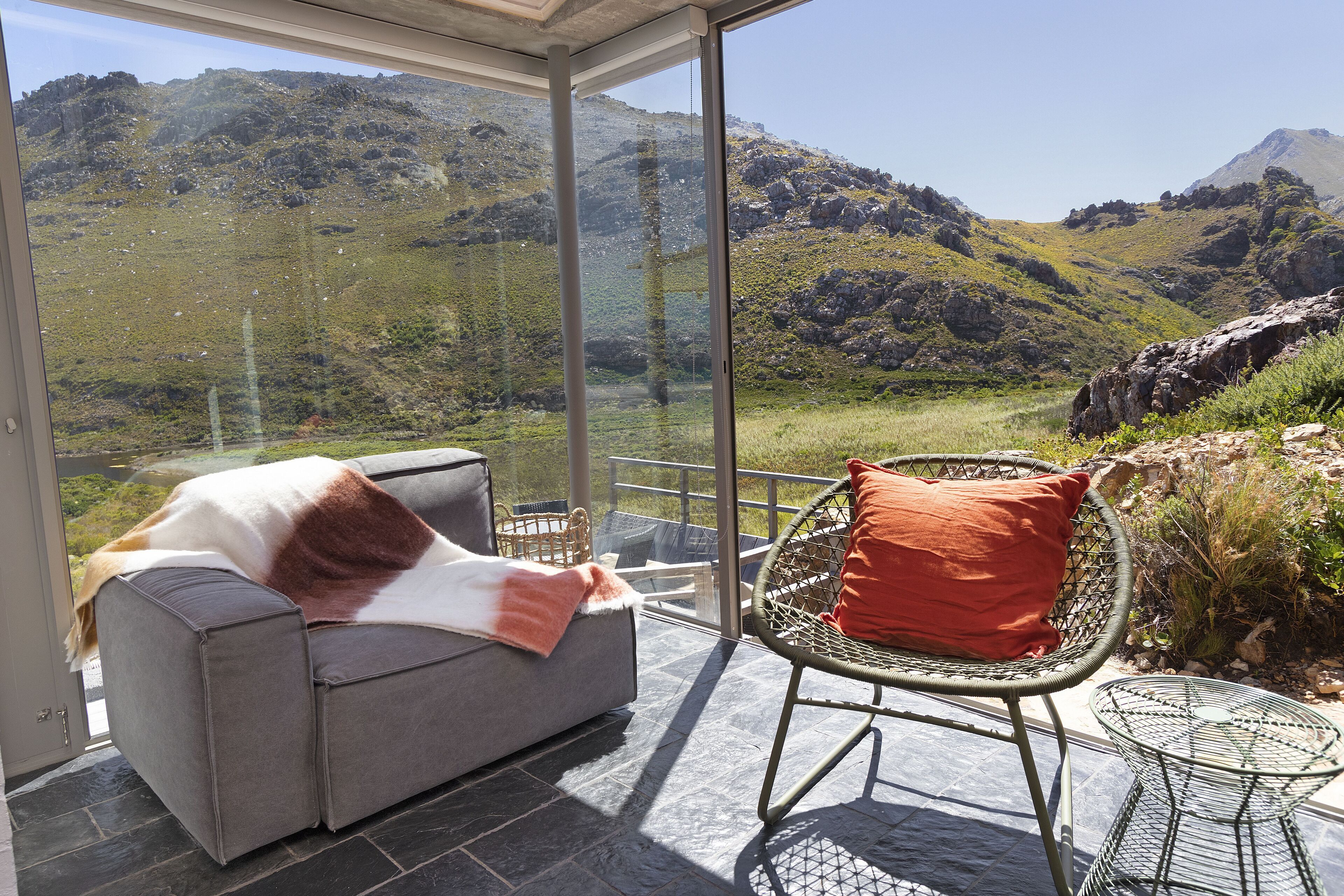 A modern room with a glass wall offering a mountain view, a grey sofa, and a wicker chair.