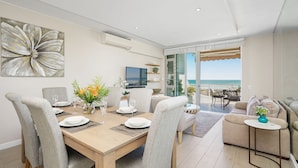 A modern living room with a dining area, a television, and a view of the beach.