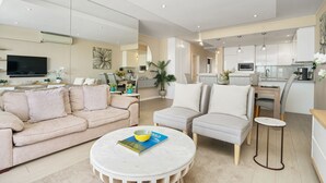 A modern living room with a sofa, armchairs, and a coffee table. There is a kitchen area with cabinets and a dining table.