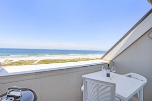 A balcony with a view of the beach, a table with a bottle and glasses, and a white chair. - Dolphin Beach H301 - One Bedroom Apartment, Sleeps 2 (Cape Town)