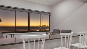 A modern room with a large window overlooking a sunset. There are three white chairs and a small table.