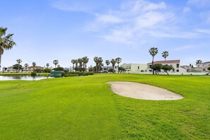 Un campo de golf con un búnker de arena, palmeras y edificios residenciales al fondo.