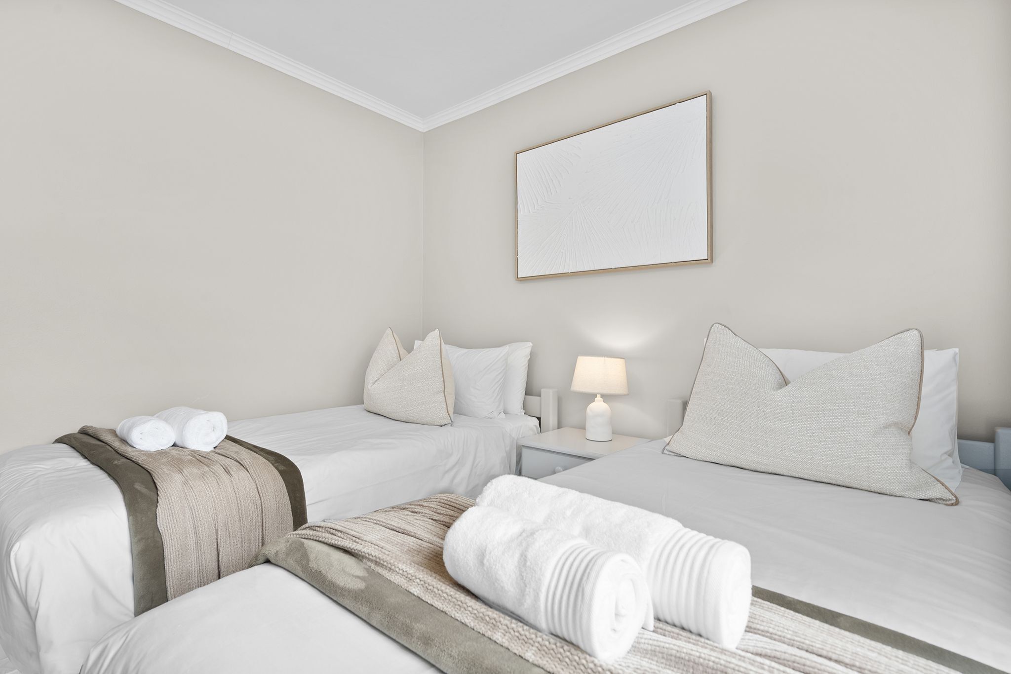 A hotel room with two beds, a bedside table with a lamp, and a wall-mounted artwork.