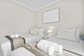 A hotel room with two beds, a bedside table with a lamp, and a wall-mounted artwork. - Laguna Beach B8 - Three Bedroom Apartment, Sleeps 6 (Cape Town)