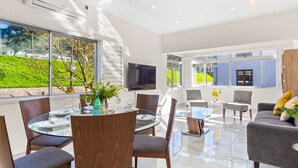 A modern living room with a glass dining table, comfortable seating, a flat-screen TV, and large windows offering a view of a green landscape.