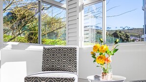 A modern living room with a patterned armchair, a small round table with a vase of yellow flowers, and a view of greenery and buildings through large windows.