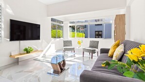 A modern living room with a flat-screen TV, glass coffee table, grey sofa, patterned chairs, and large windows offering a view of greenery.