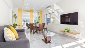 A modern living room with a sofa, dining table, and chairs. There is a TV mounted on the wall and a plant on a shelf.