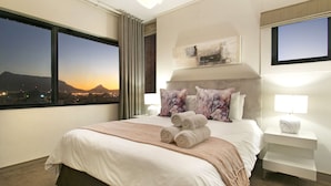 Ein Hotelzimmer mit einem großen Bett, Blick auf eine Stadtlandschaft im Sonnenuntergang, einem Flachbildfernseher und einem Nachttisch mit Lampe.