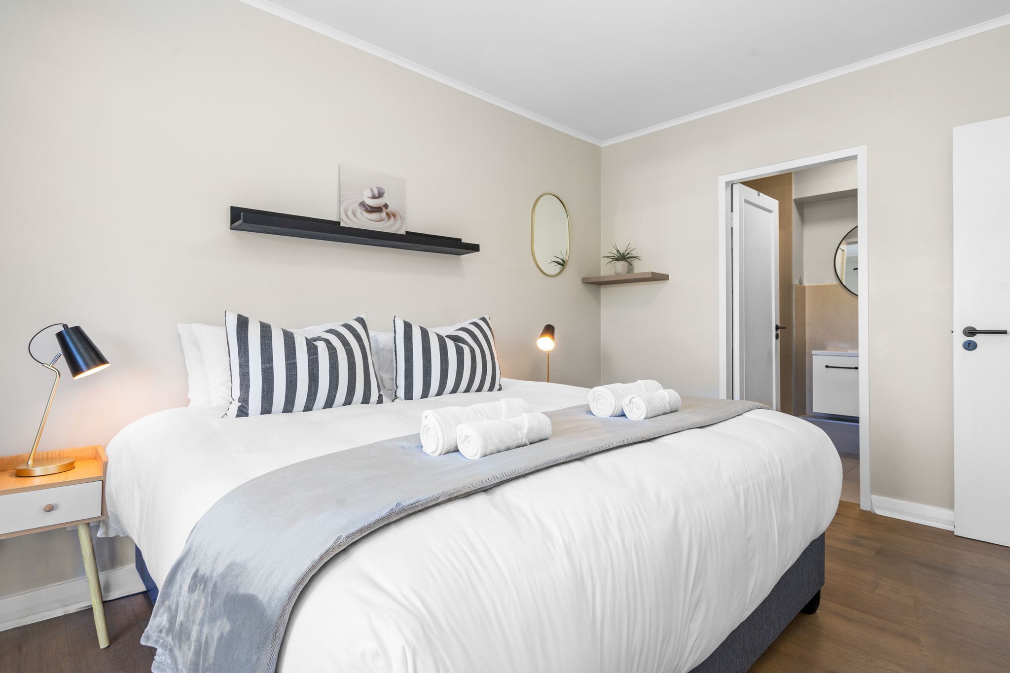 Una cama bien tendida con ropa de cama blanca y almohadas a rayas, una lámpara de noche y un pequeño estante con adornos.