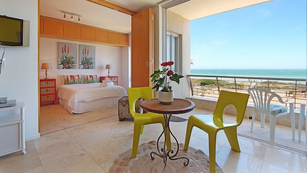 A hotel room with a bed, a small table with chairs, a TV, and a balcony with a view of the sea. - Neptune Isle 214 - One Bedroom Apartment, Sleeps 2 (Cape Town)