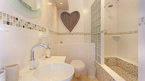 A bathroom with a large white sink, a toilet, a shower with mosaic tiles, and a heart-shaped decoration on the wall. - Neptune Isle 214 - One Bedroom Apartment, Sleeps 2 (Cape Town)