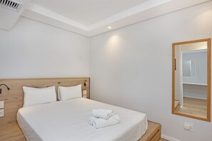 Cama bem arrumada com roupa de cama branca e toalhas dobradas, cabeceira de madeira e um espelho grande na parede.