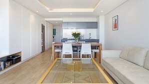 Una sala moderna con una mesa de comedor de vidrio, sillas blancas, un sofá color beige y una cocina al fondo.
