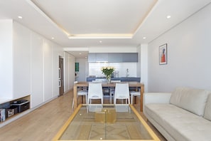 A modern living room with a glass dining table, white chairs, a beige sofa, and a kitchen in the background.