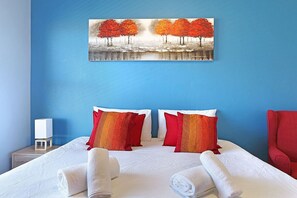 A hotel room with a bed, two pillows, a red chair, a painting of trees, and a nightstand with a lamp.