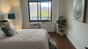 Quarto com uma cama grande, janela com vista para edifícios, radiador e uma planta em vaso.