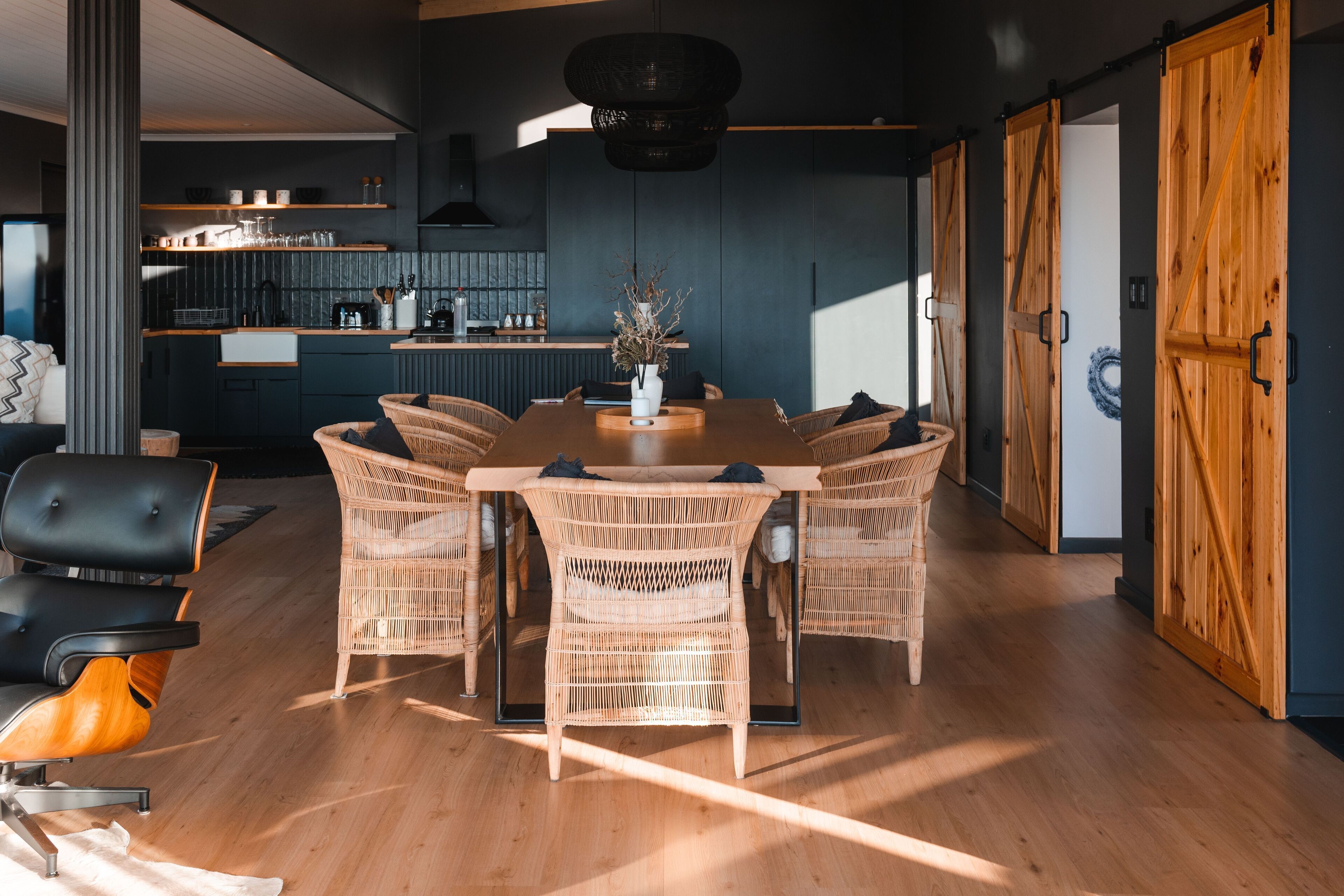Een moderne eethoek met een houten tafel en rieten stoelen, een keuken op de achtergrond en een grote houten deur.