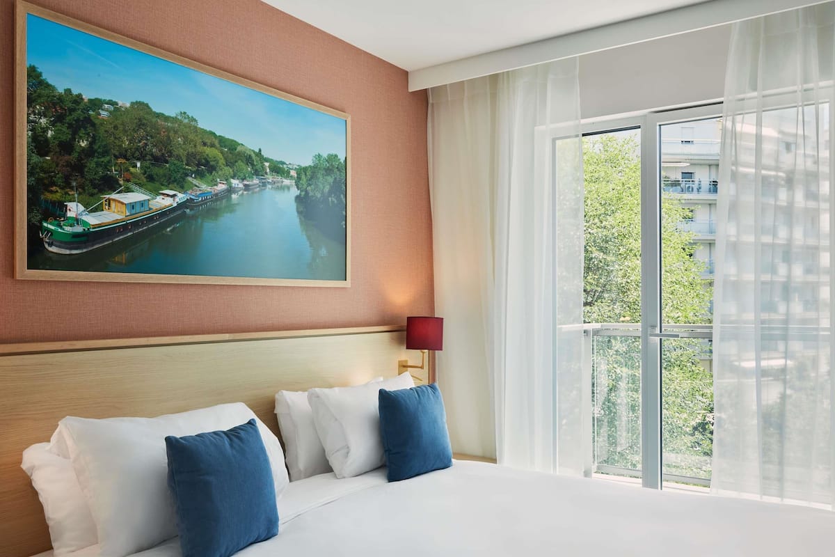 Une chambre d’hôtel avec un grand lit, un tableau représentant une rivière et une vue sur la verdure par la fenêtre.