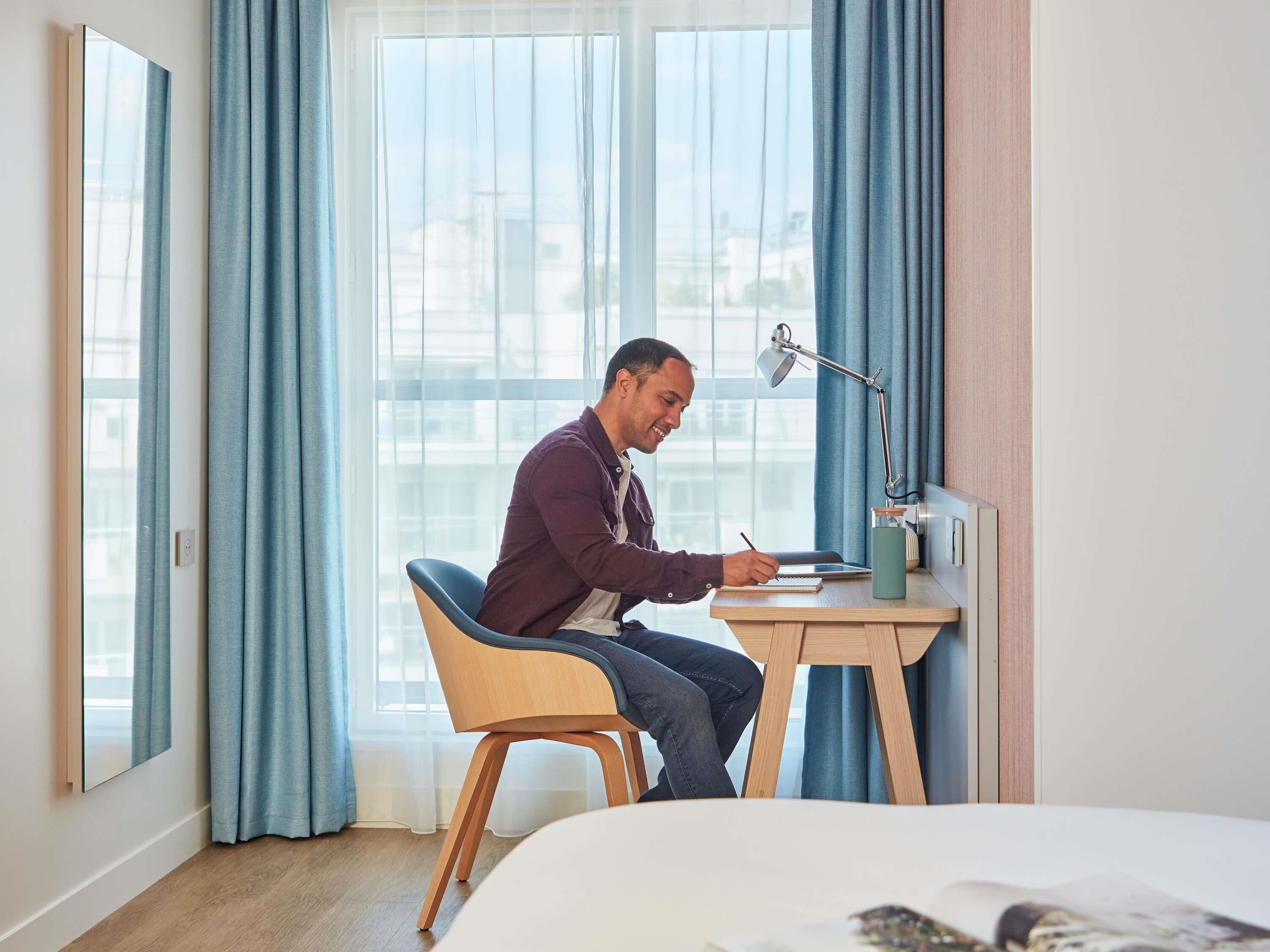 A man sitting at a desk in a hotel room with a laptop, writing. There is a lamp on the desk, and a bed with a magazine on it.