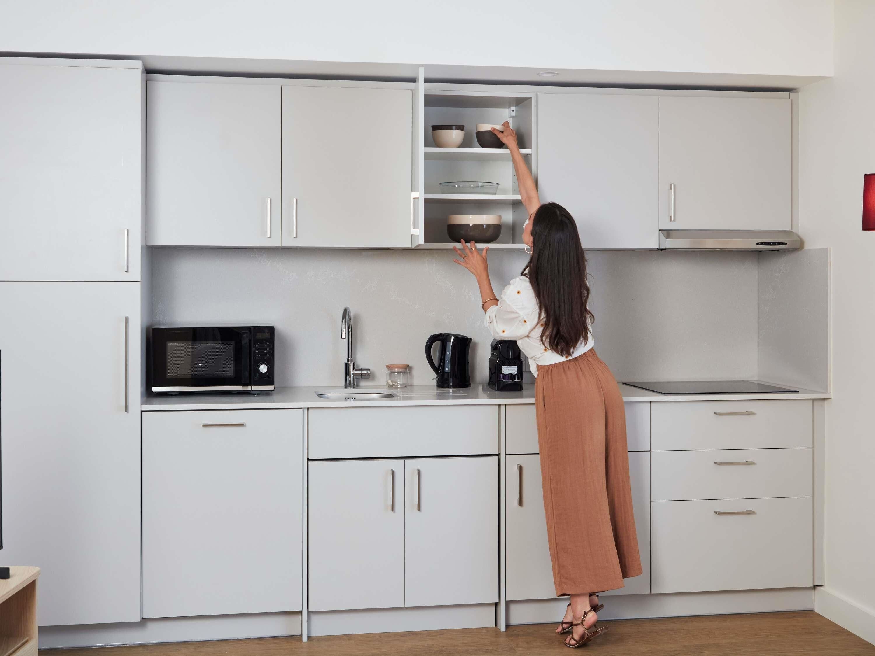 Una cocina moderna con armarios blancos, un microondas, una cafetera y una persona alcanzando objetos en el estante superior.