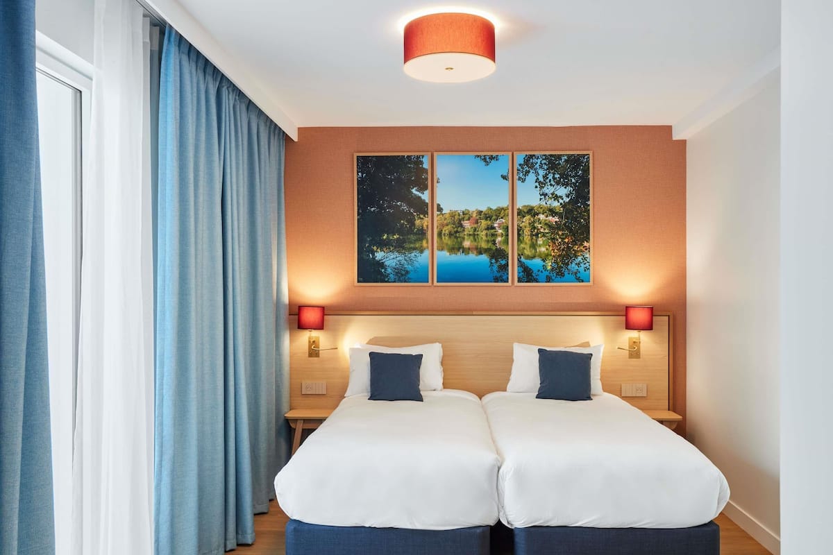 Cama doble con dos almohadas, una mesita de noche, una lámpara de pared y un cuadro sobre la cama.