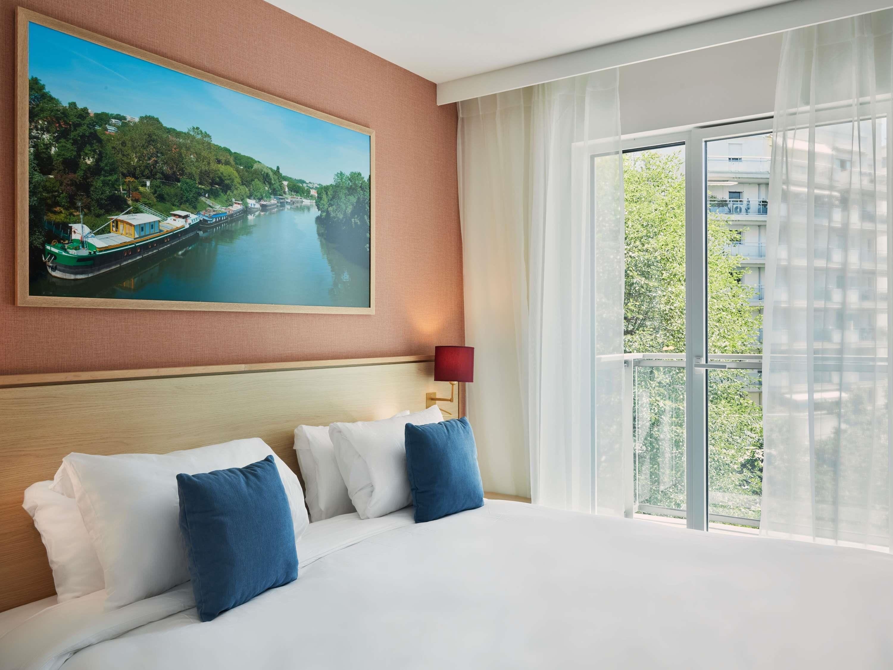 Ein Hotelzimmer mit einem großen Bett, einem Gemälde einer Flusslandschaft, einem Fenster mit Vorhängen und einem Nachttisch mit Lampe.
