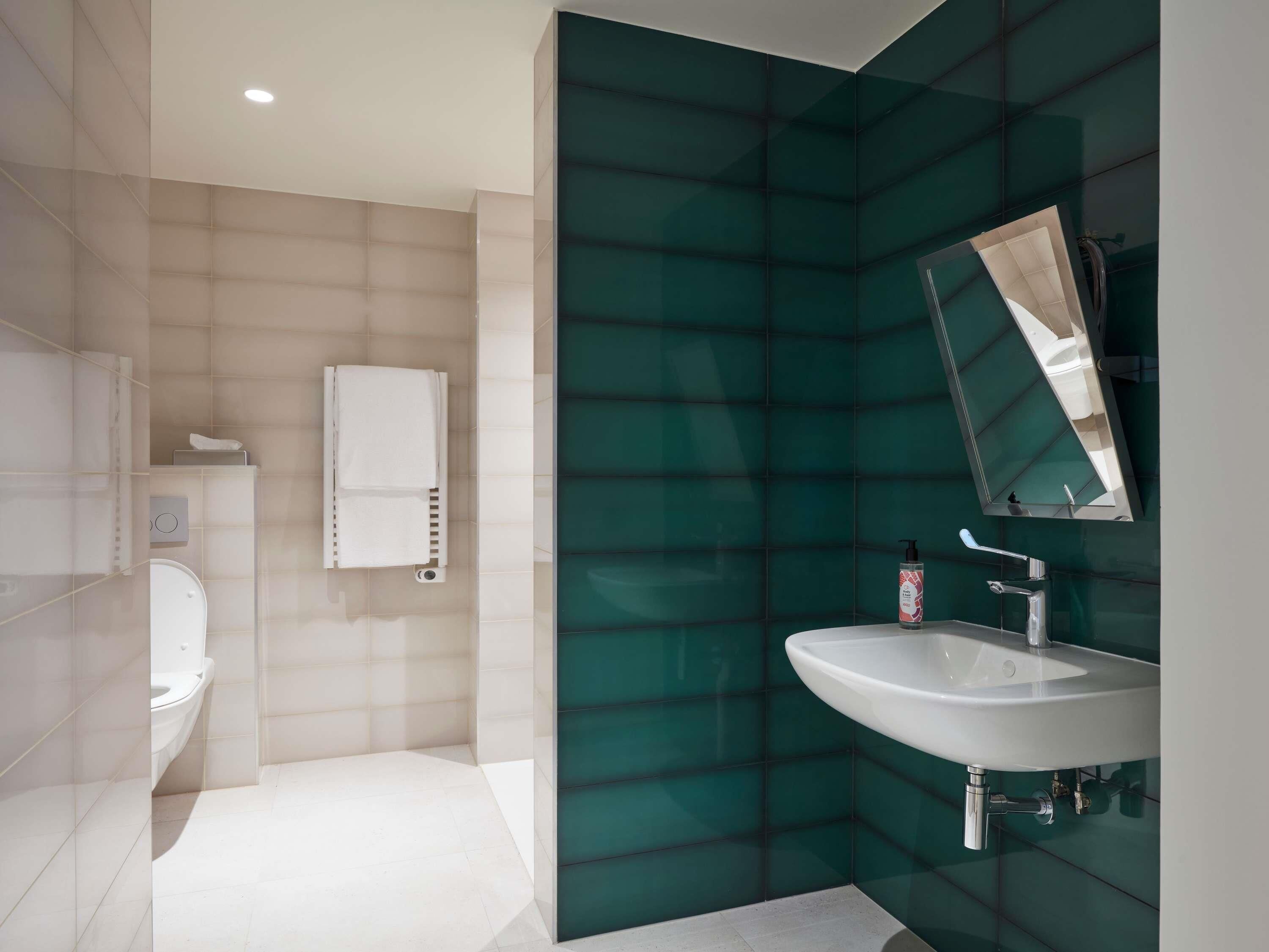 Uma casa de banho moderna com sanita branca, lavatório branco e parede revestida a azulejos verdes.