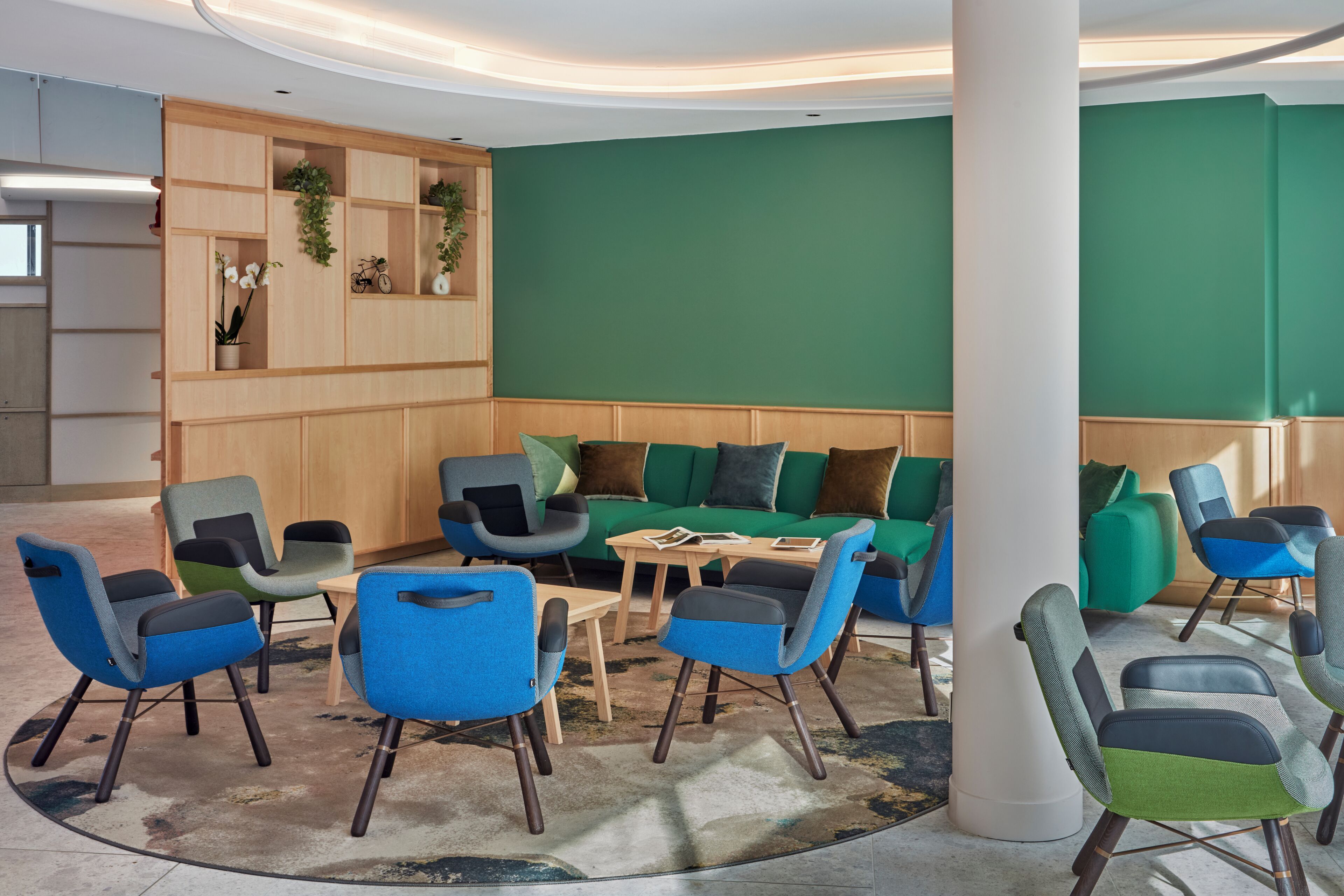 A modern lounge area with a green wall, wooden shelving, and blue and grey upholstered chairs.