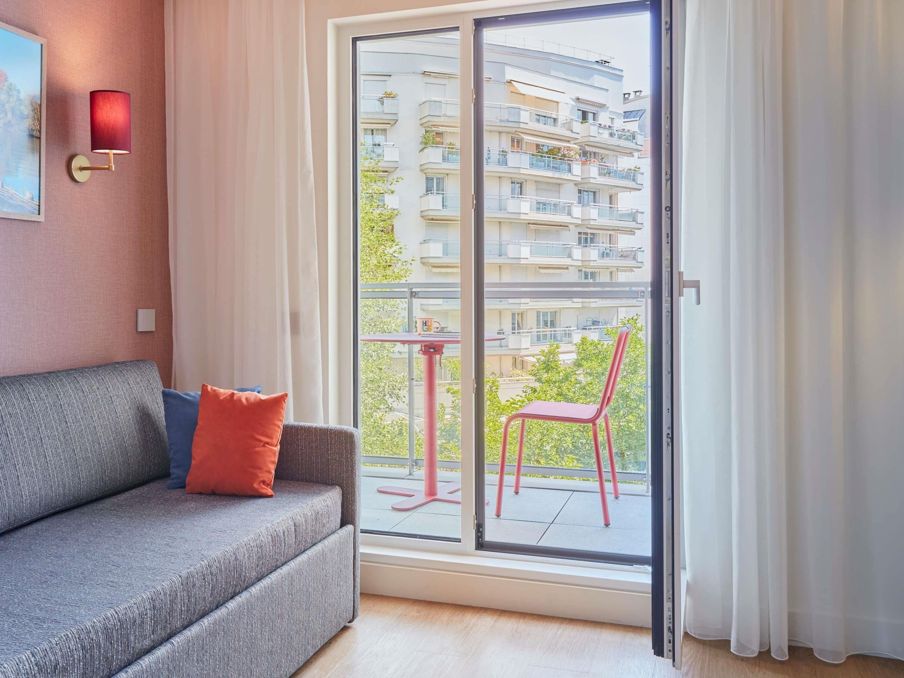 Un salon moderne avec un canapé gris, un fauteuil rouge et une vue sur un immeuble à plusieurs étages, visible à travers une porte vitrée.