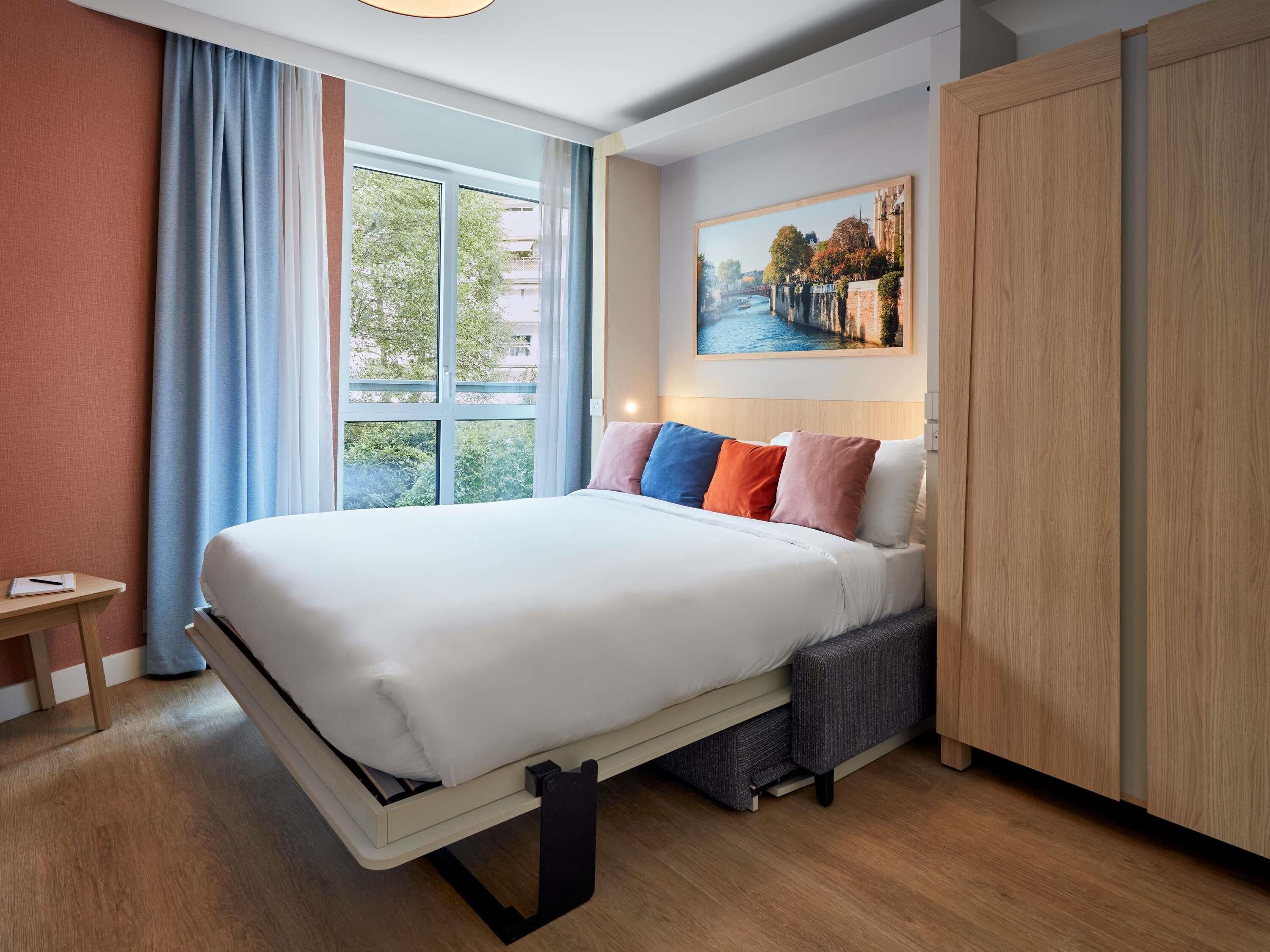 A hotel room with a bed, a desk, a window with curtains, and a painting on the wall.