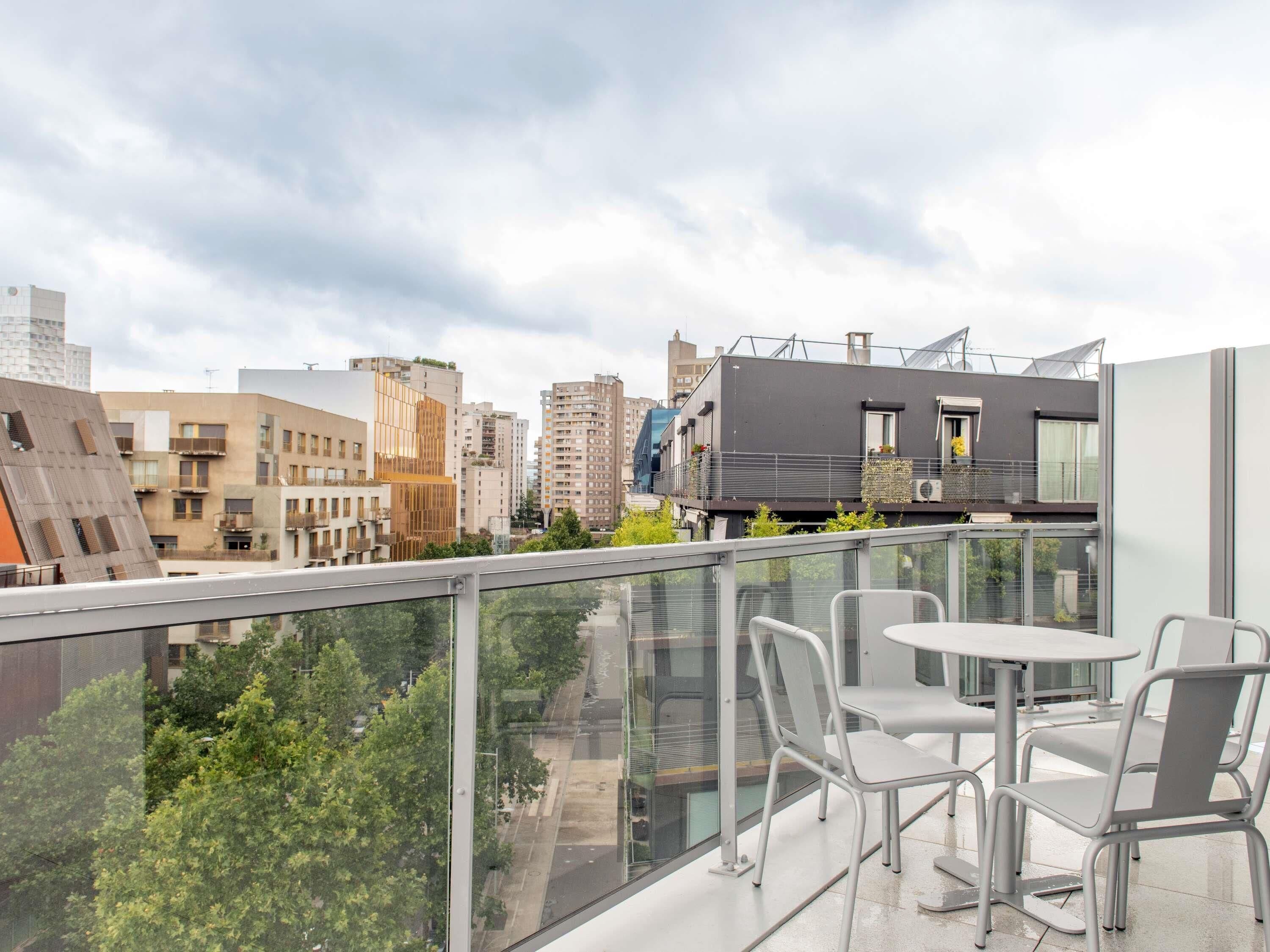 Un balcón moderno con barandillas de vidrio, una mesa y sillas, con vista a un paisaje urbano con varios edificios.