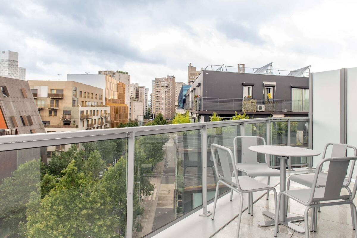 Un balcon moderne avec des garde-corps en verre, une table et des chaises, offrant une vue sur un paysage urbain composé de différents bâtiments.