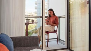 Eine Person sitzt auf einem roten Stuhl auf einem Balkon mit Blick auf Gebäude und einer Pflanze auf einem Tisch.