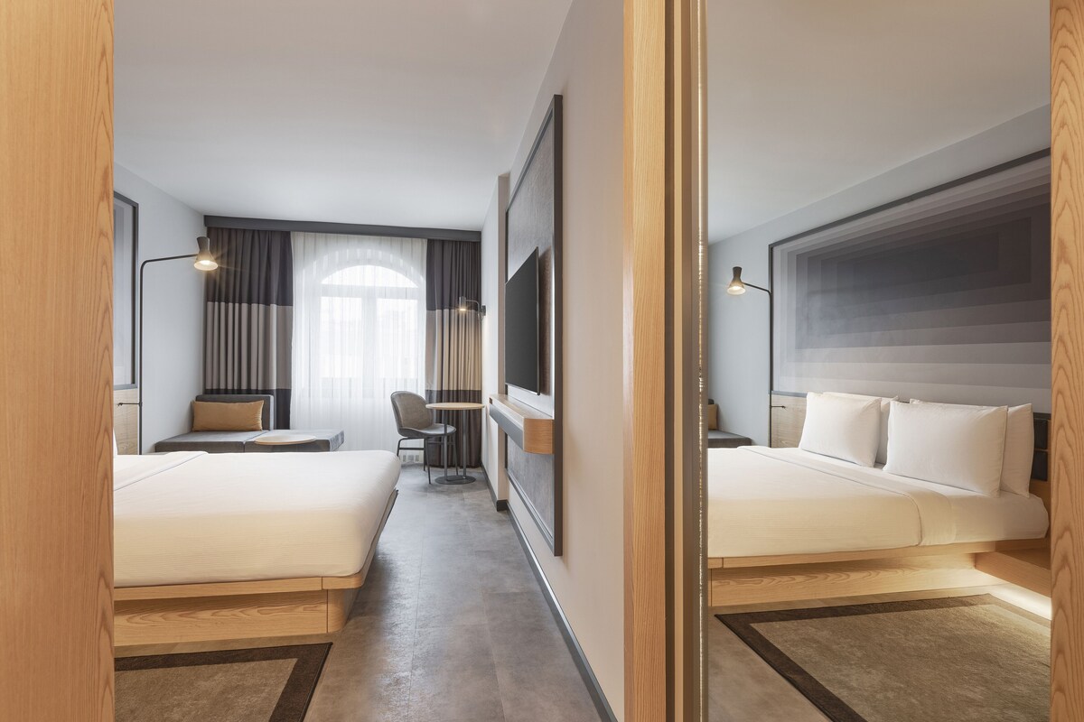 A hotel room with two beds, a desk, a chair, a TV, and a large window with curtains.