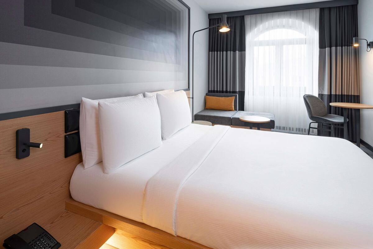 A hotel room with a large bed, bedside controls, a desk, a chair, and a window with curtains.