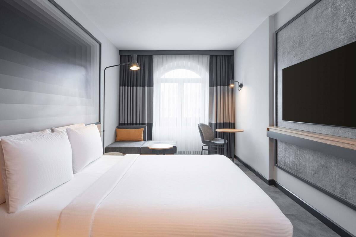 A modern hotel room with a large bed, a small seating area, a desk, and a large window with curtains.