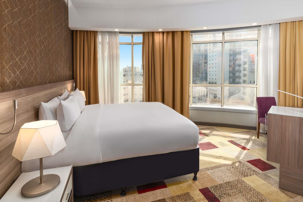 A hotel room with a large bed, a desk with a lamp, a chair, and a view of the city through large windows.