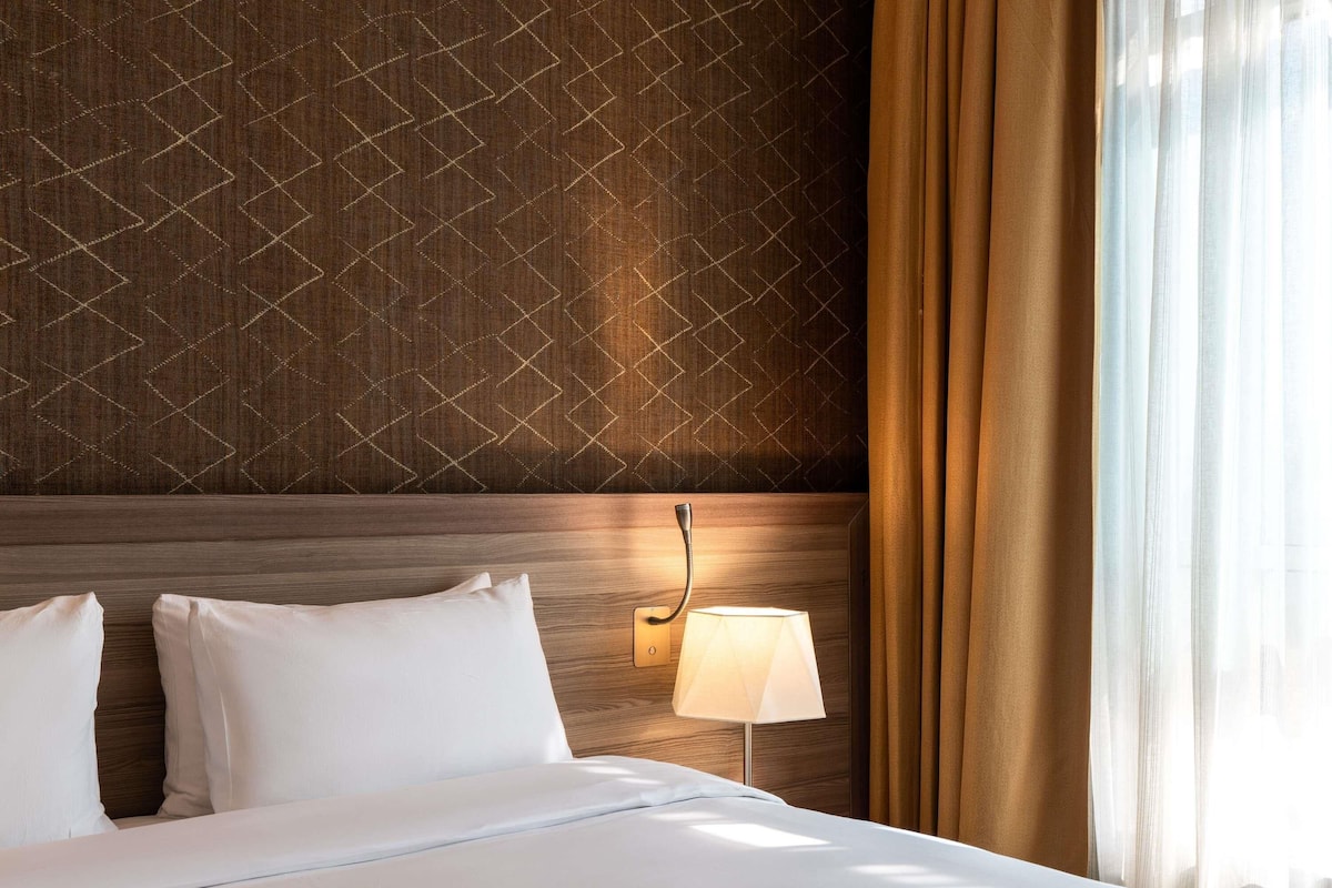 Una habitación de hotel con un cabecero estampado, una lámpara de noche y una ventana con cortinas.